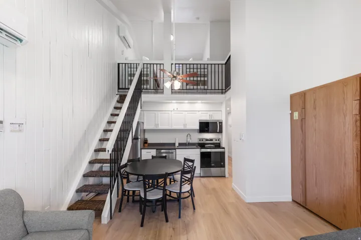 Open-concept living space with wood-finish flooring, a contemporary kitchen featuring stainless steel appliances and white cabinetry, a built-in wooden cabinet, and a loft area with a ceiling fan