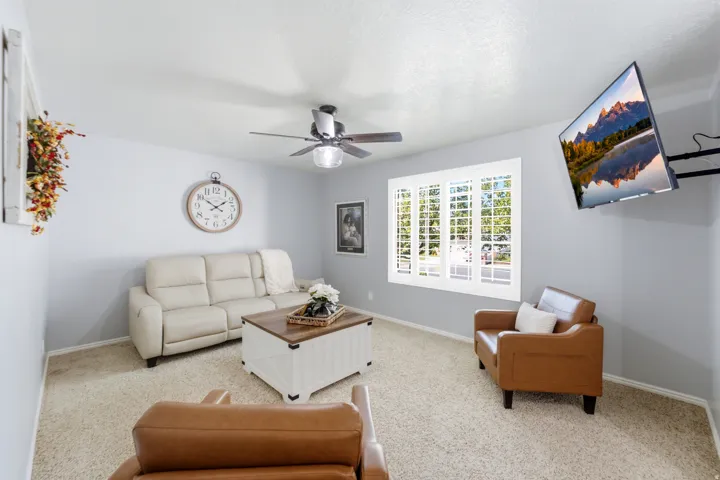 Carpeted living area featuring a ceiling fan with light fixture, window with white plantation shutters, and light gray wall paint