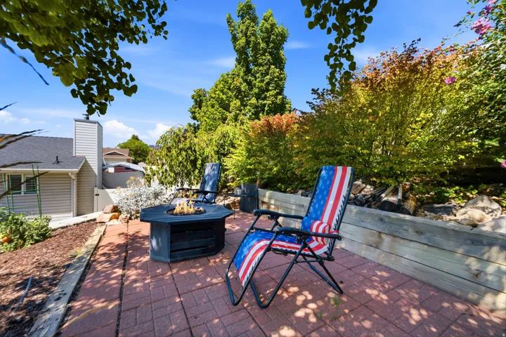 Brick paver patio featuring a built-in fire pit, surrounded by lush landscaping and a timber retaining wall