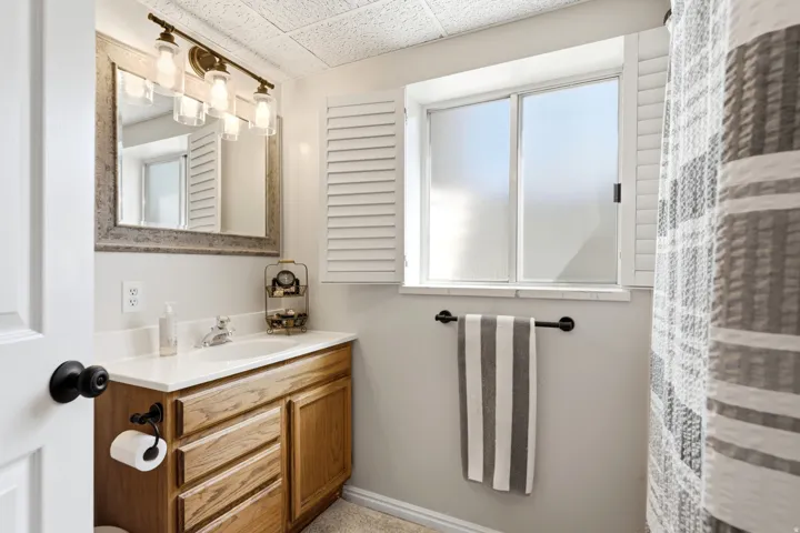 Bathroom featuring a wood-finish vanity with a single sink, light-colored countertop, framed mirror, and horizontal window with privacy shutters