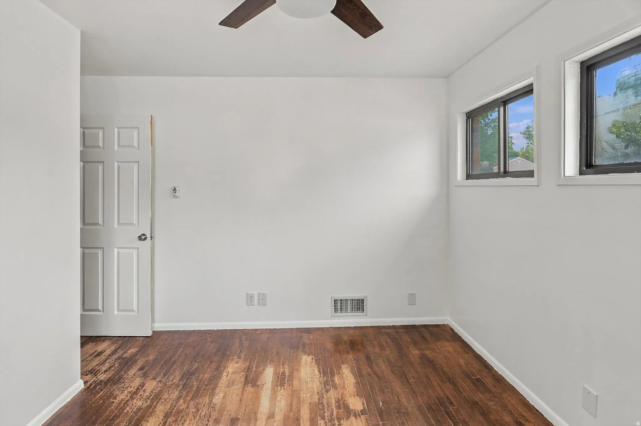 Wood-finish flooring, white paneled door, two windows with dark frames, and a ceiling fan with wood-tone blades