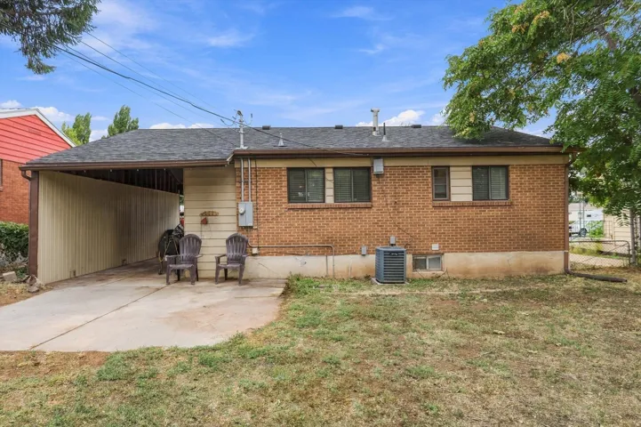 Brick exterior with a carport and concrete patio, featuring a dark shingle roof and mature trees