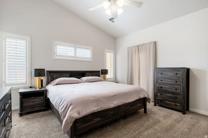 Vaulted ceiling bedroom with plantation shutters, ceiling fan with light, and light-colored carpeting