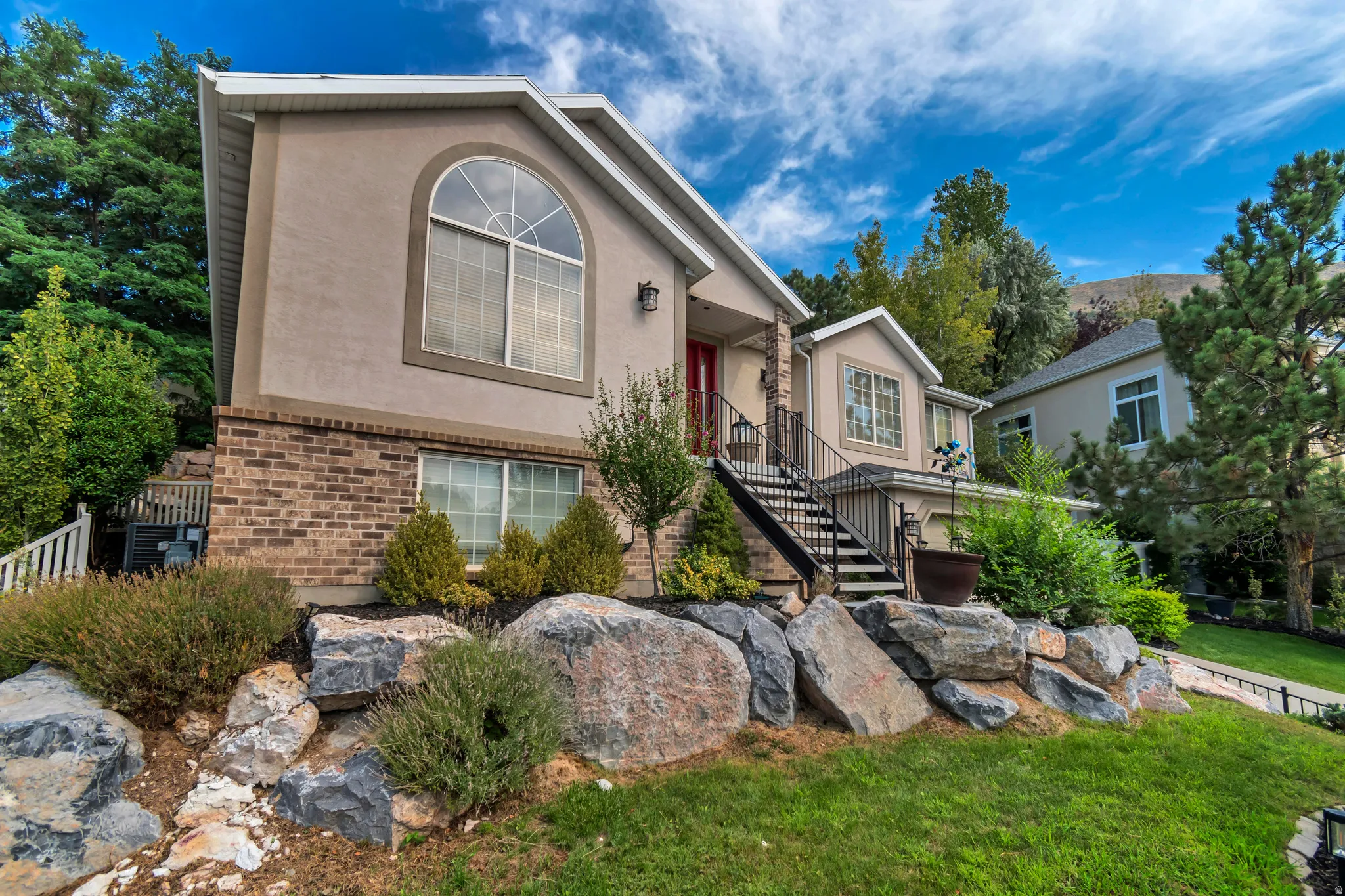 Stucco and brick exterior with an arched window, complemented by a tiered rock garden, established landscaping, and a red entry door