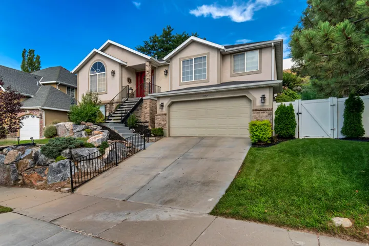 Stucco exterior with brick accents, prominent two-car garage, established front landscaping with rock features, and a white privacy fence