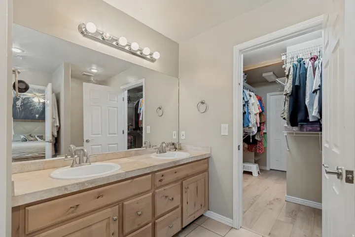 Dual vanity with integrated sinks, stone-finish countertop, light wood cabinetry, and an adjacent walk-in closet with built-in shelving