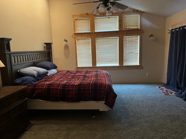 Vaulted ceiling with a ceiling fan, multiple windows with natural wood trim, wall-mounted light fixtures, and carpeting