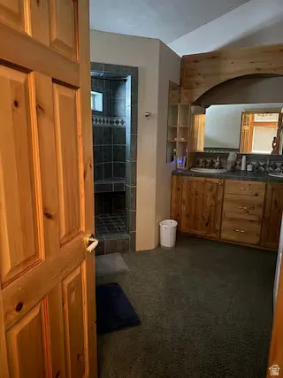 Wood panel door entry to a bathroom featuring a tile-surround shower, wood-finish vanity with dual sinks, and an arched wood beam accent