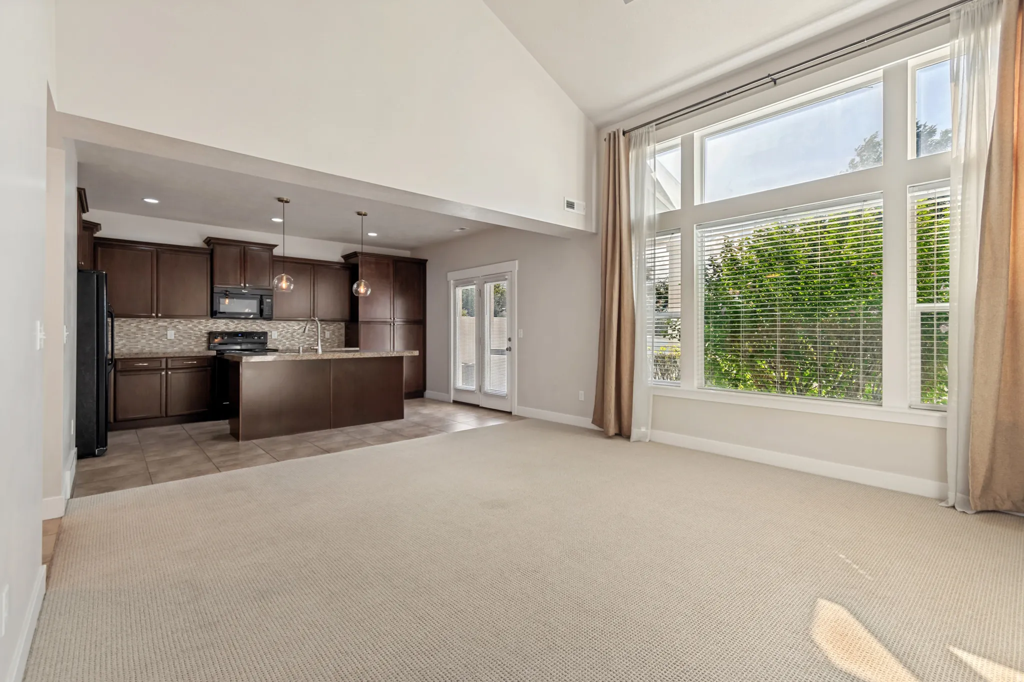 Vaulted ceiling, expansive windows with blinds, carpeted flooring, kitchen with dark wood-finish cabinetry, and a center island with pendant lighting