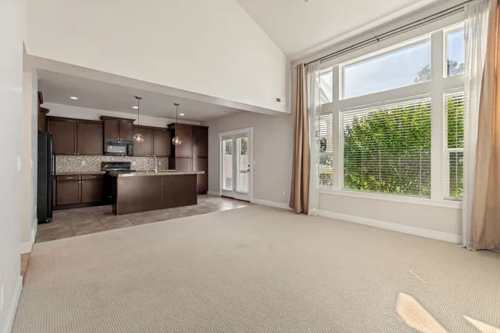 Vaulted ceiling, expansive windows with blinds, carpeted flooring, kitchen with dark wood-finish cabinetry, and a center island with pendant lighting