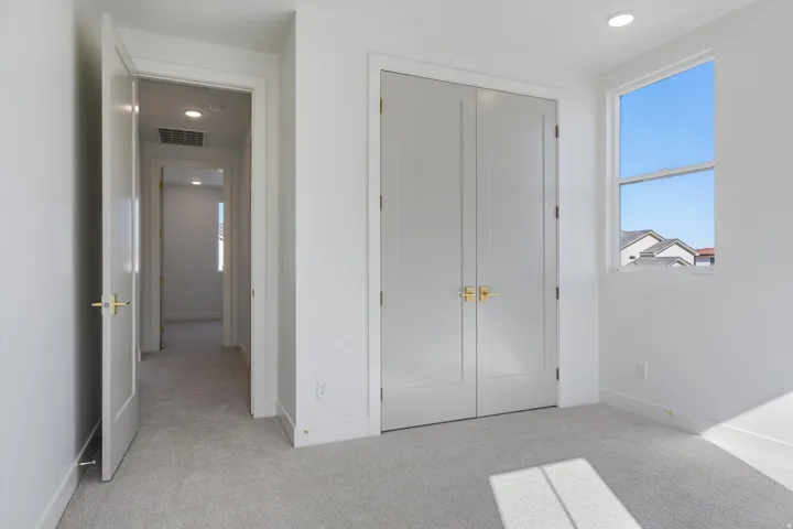 This bedroom has plush carpet flooring, light walls, double-door closet with gold-finish hardware, recessed lighting, and a three windows