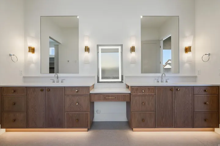 Double vanity with white countertops, wood-finish cabinetry, and contemporary wall sconces and lighted mirrors