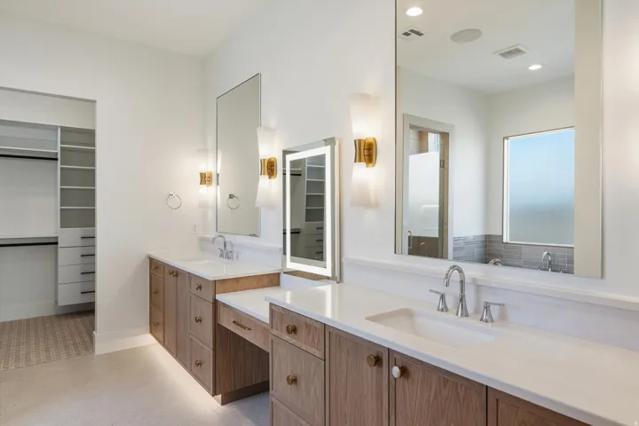 Double vanity with white countertops, wood-finish cabinetry, and contemporary wall sconces and lighted mirrors
