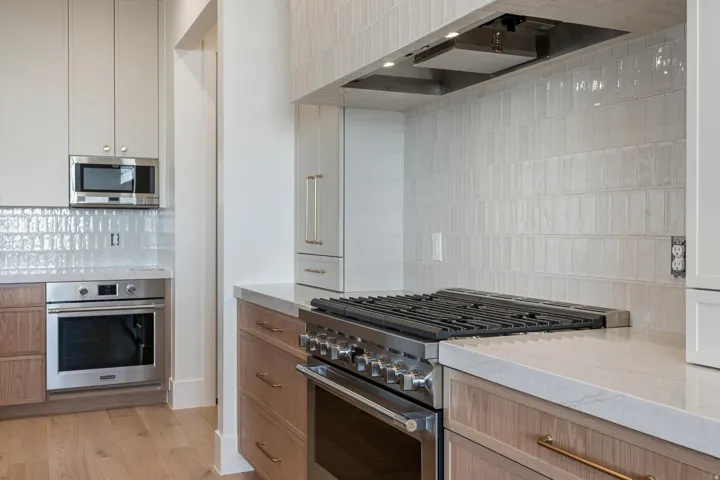 Stainless steel gas range, integrated microwave, wood-finish lower cabinetry, white upper cabinetry, and a full-height tile backsplash