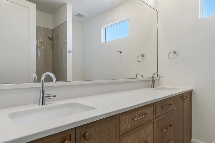 Dual vanity with integrated sinks, chrome fixtures, a full-width mirror, wood-finish cabinetry, and a walk-in shower with tiled walls