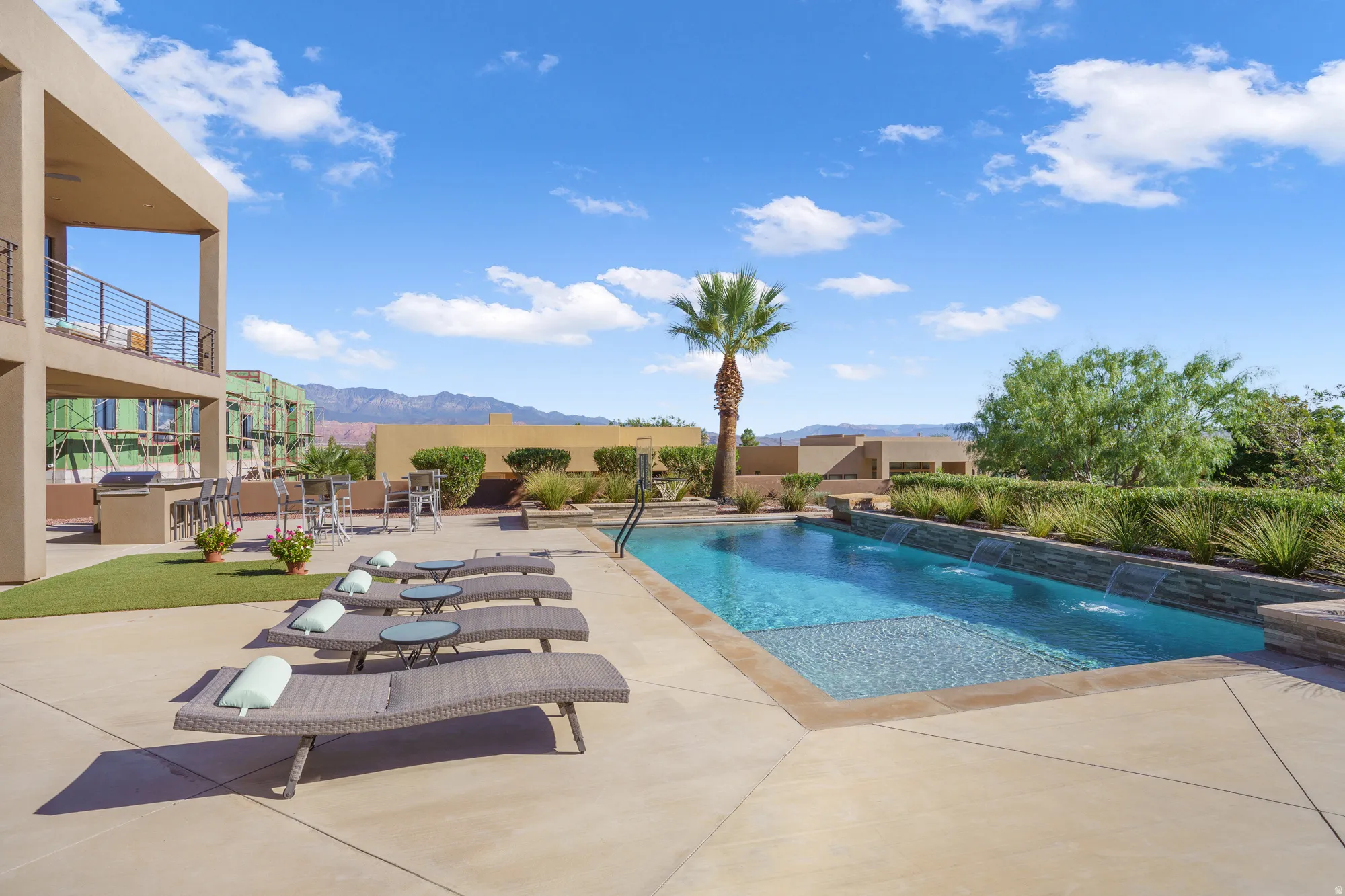 Paved patio area, rectangular swimming pool with water features, integrated outdoor kitchen, and an upper-level balcony with metal railing