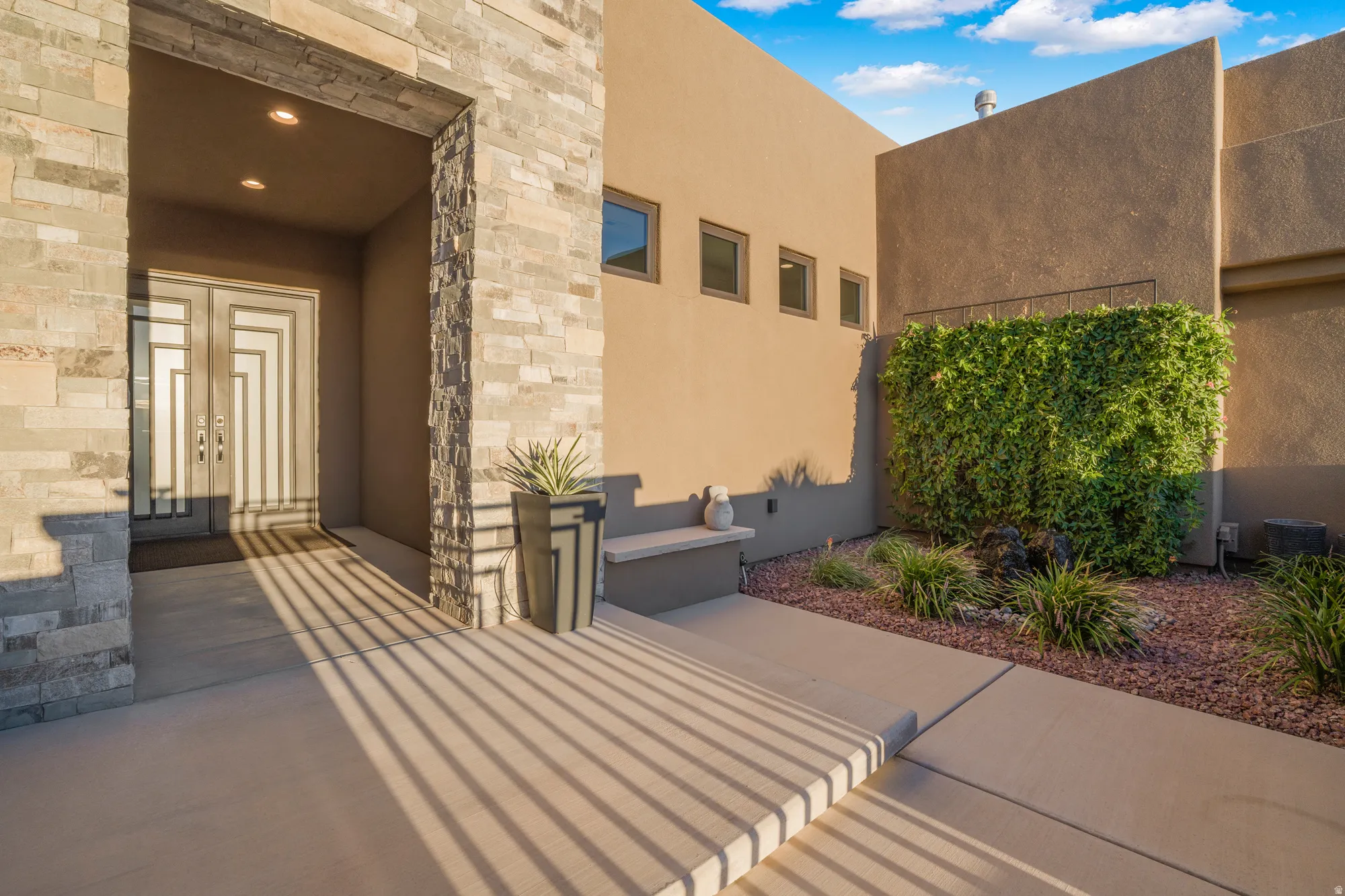 Stucco exterior featuring stacked stone accents, a double entry door with geometric detailing, recessed lighting, and a landscaped courtyard with decorative shrubs