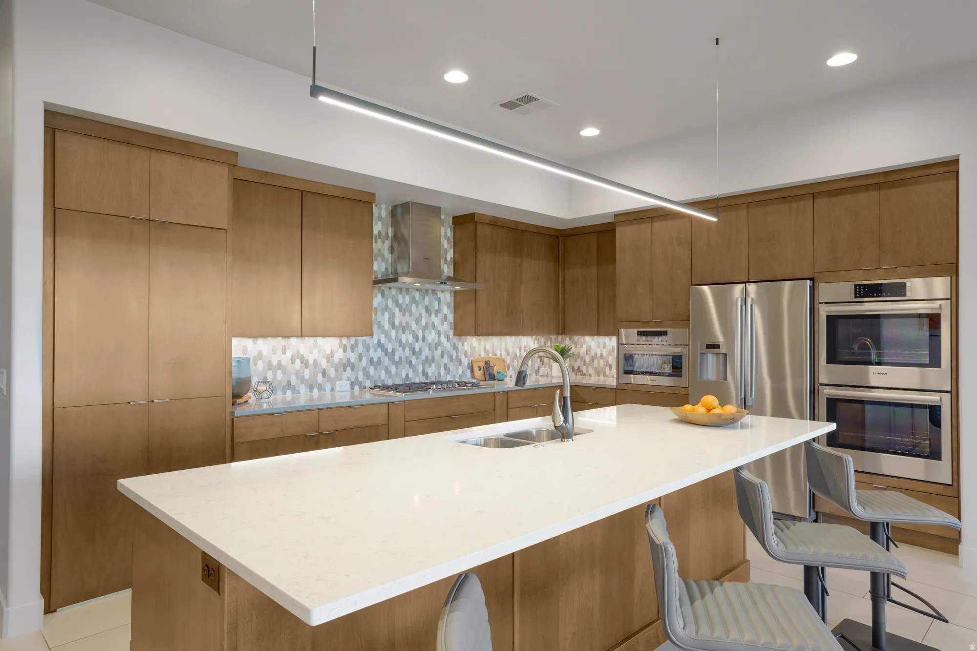 Kitchen featuring light wood-finish cabinetry, a white countertop island with an integrated sink, stainless steel appliances, a mosaic tile backsplash, and recessed lighting