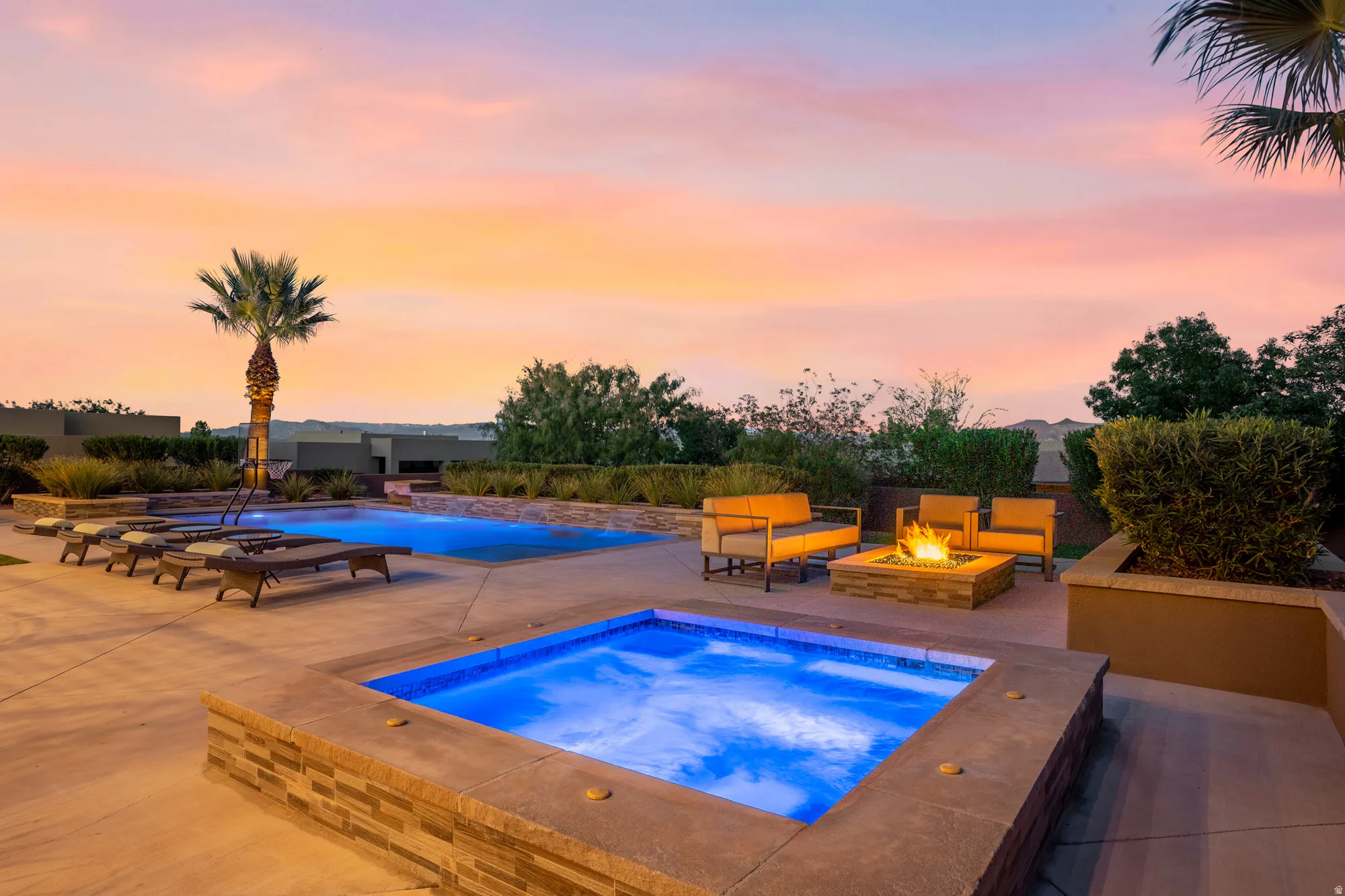 Expansive concrete patio with an illuminated swimming pool, separate spa, and stone-clad fire pit
