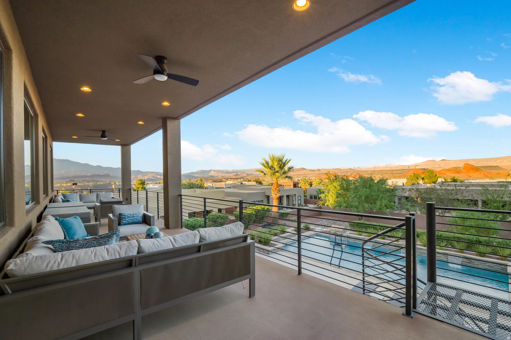 Expansive outdoor balcony with recessed lighting, ceiling fans, and a metal railing system overlooking a swimming pool and distant mountains