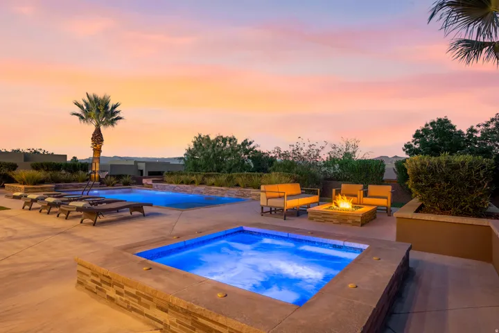 Expansive concrete patio with an illuminated swimming pool, separate spa, and stone-clad fire pit