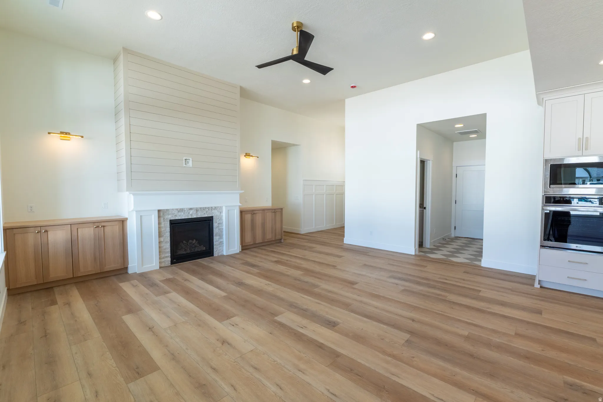 Wood-finish flooring throughout, shiplap fireplace surround with tile accent, built-in cabinetry, modern ceiling fan, and integrated wall oven and microwave