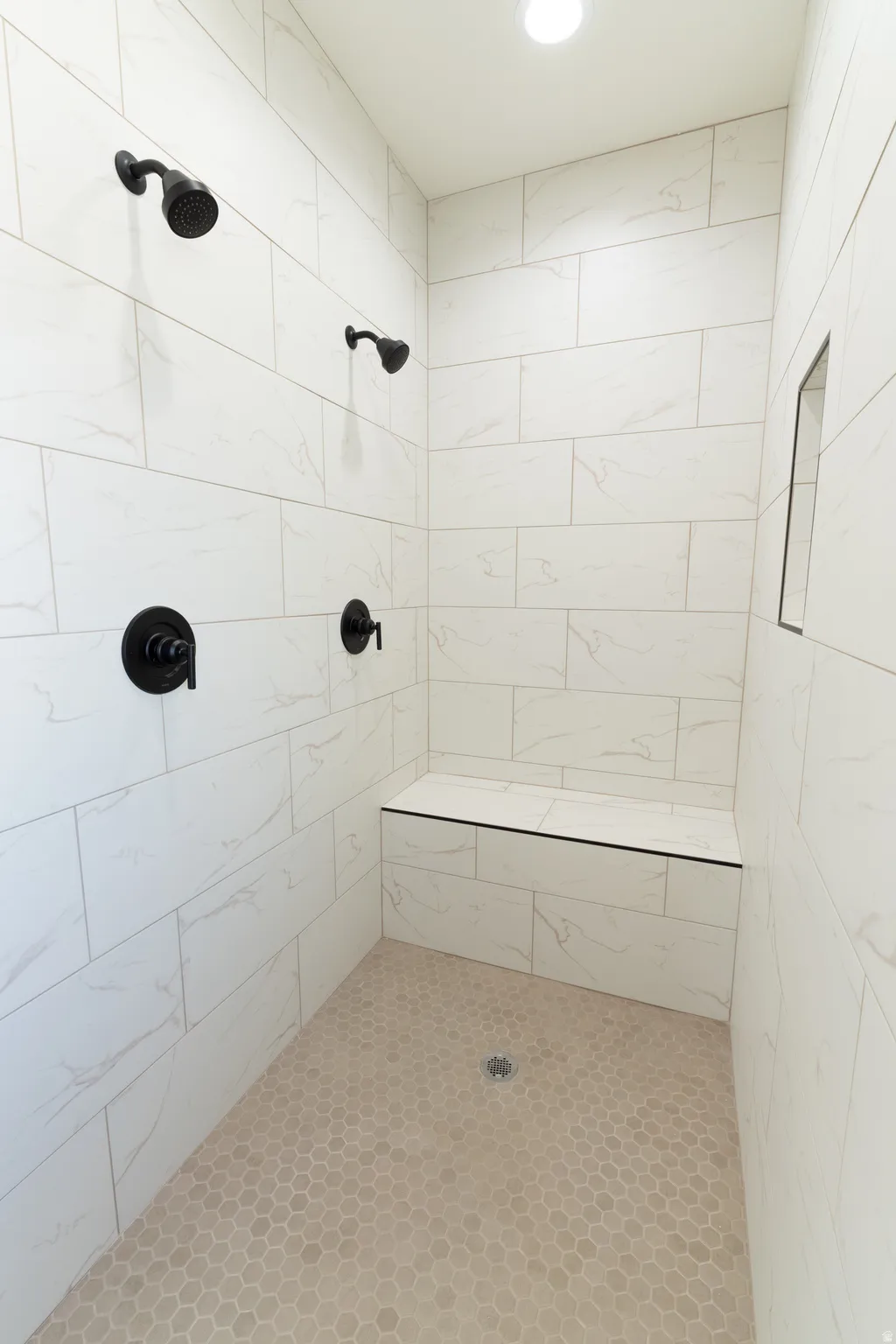 Spacious shower with large format wall tiling, two shower heads, matte black fixtures, an integrated bench, and mosaic tile flooring
