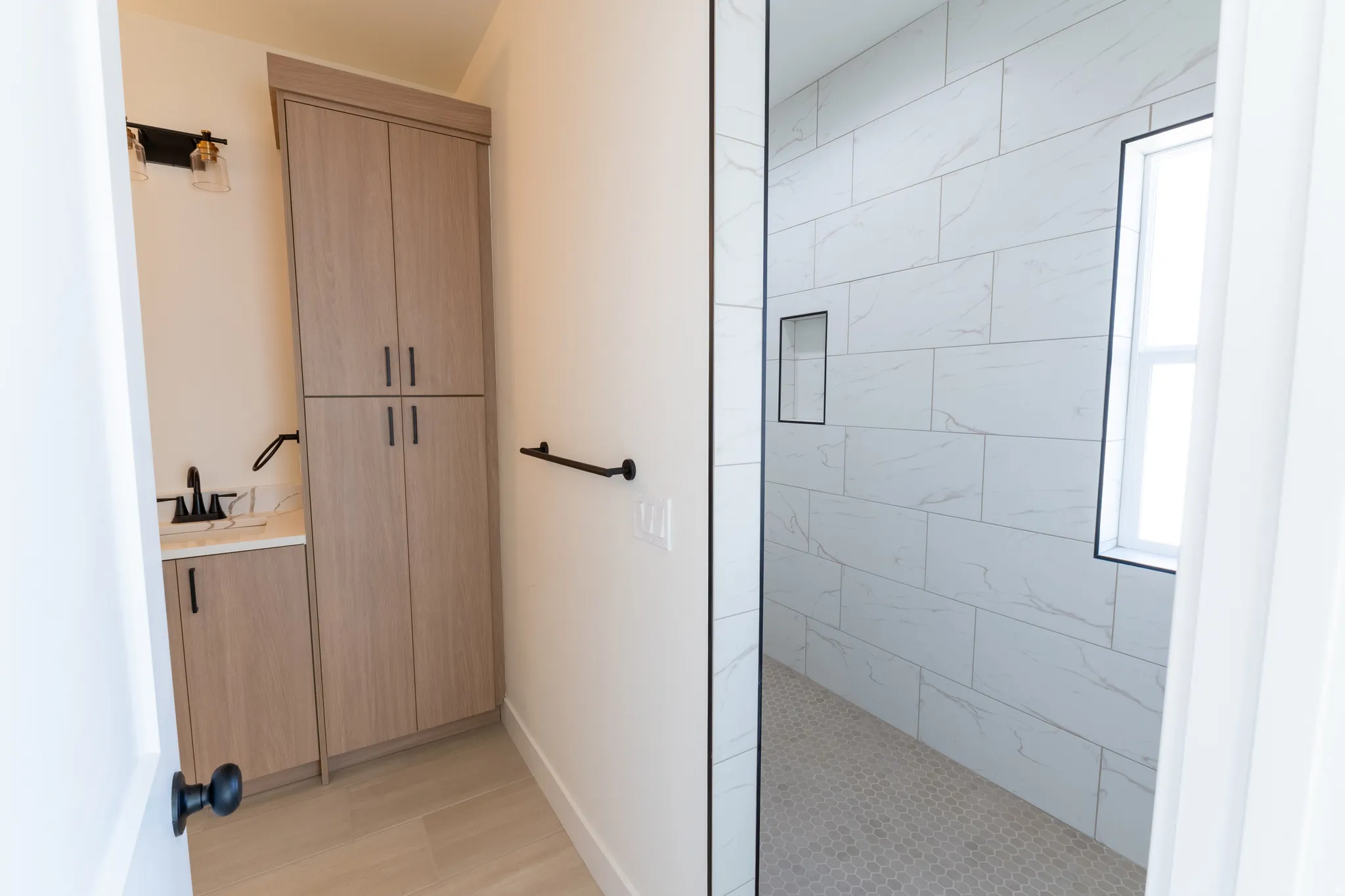 Light wood-finish cabinetry, white countertop, matte black fixtures, white tiled shower, and a shower window. Big walk-in shower.