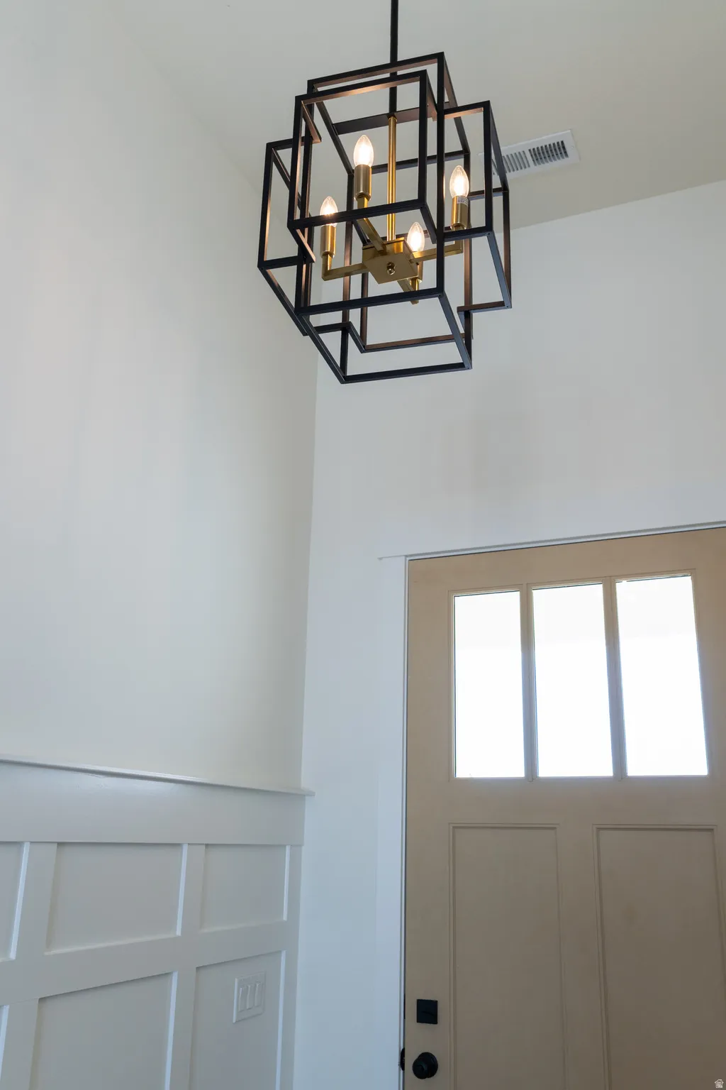 Modern geometric chandelier with brass accents, tall ceilings, board and batten wall paneling, and a light-colored entry door with glass inserts