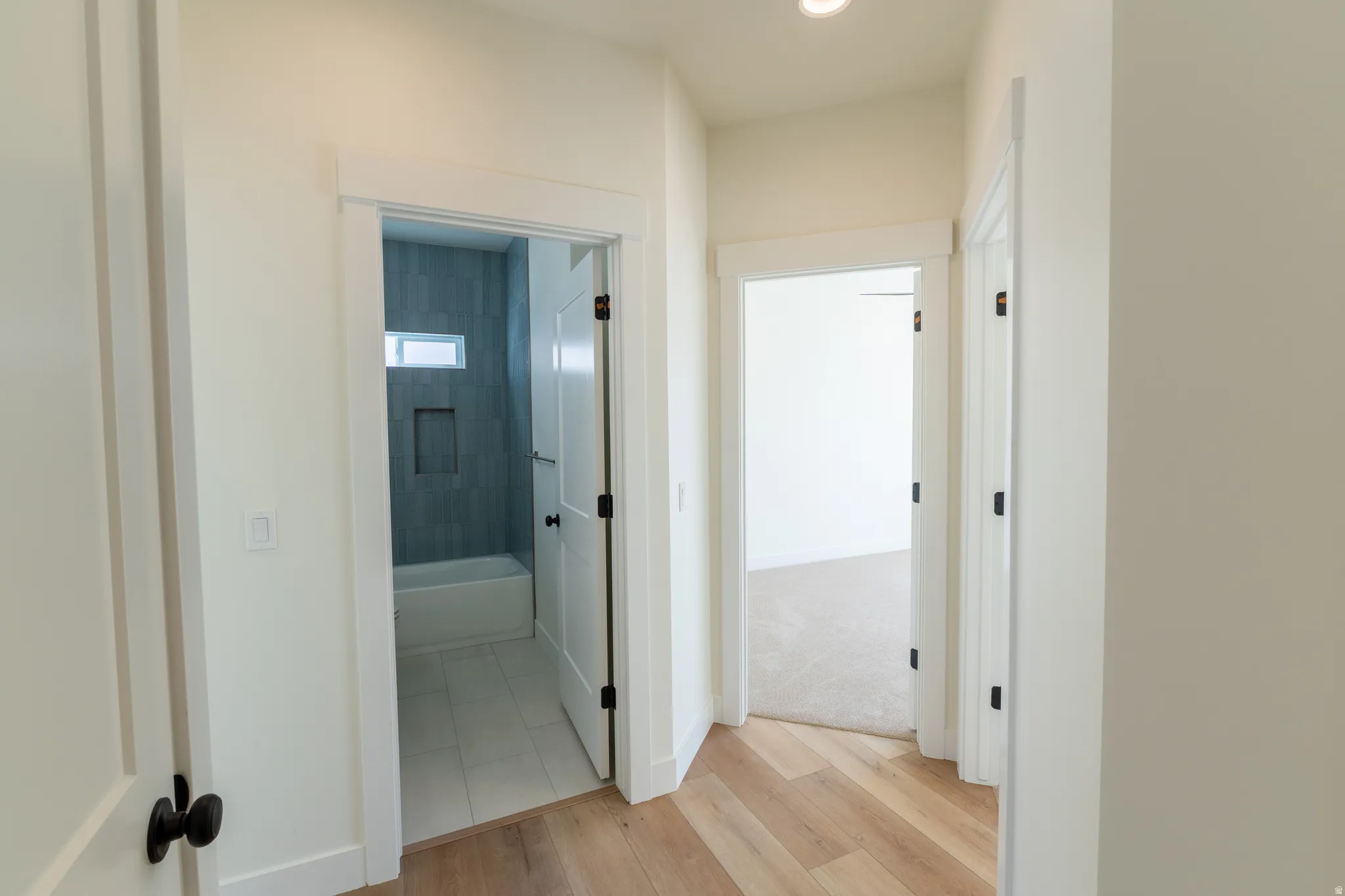 Light wood-finish flooring, white paneled doors with matte black hardware, recessed lighting, and a bathroom featuring a tub with tiled surround and an inset shelf