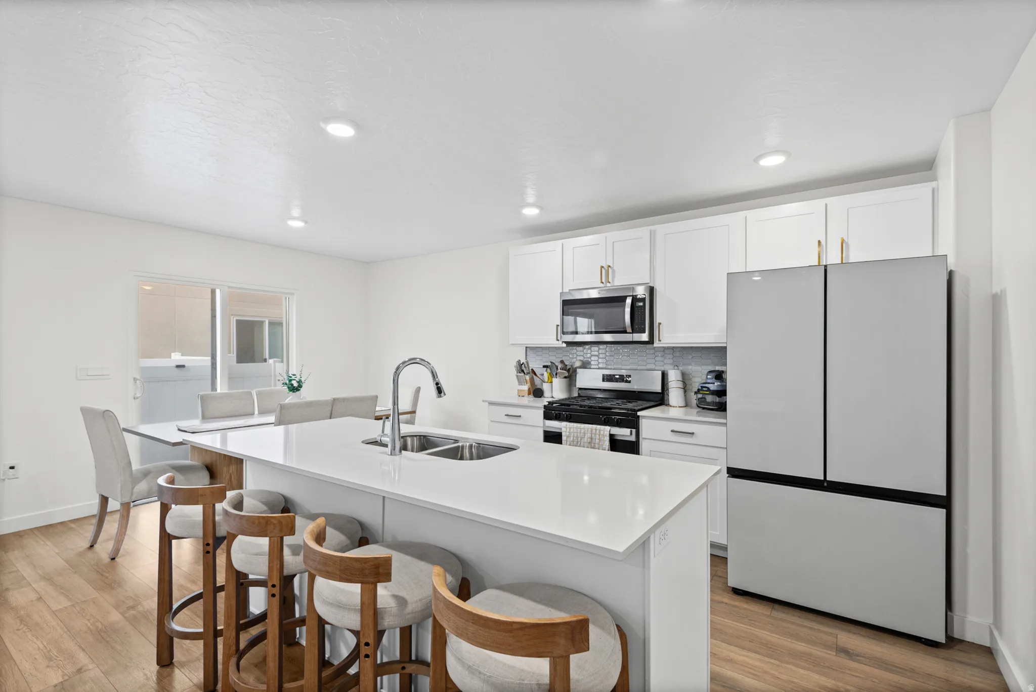 Kitchen featuring wood-finish flooring, white cabinetry with gold-tone hardware, stainless steel appliances, a tile backsplash, and a large central island with a double sink