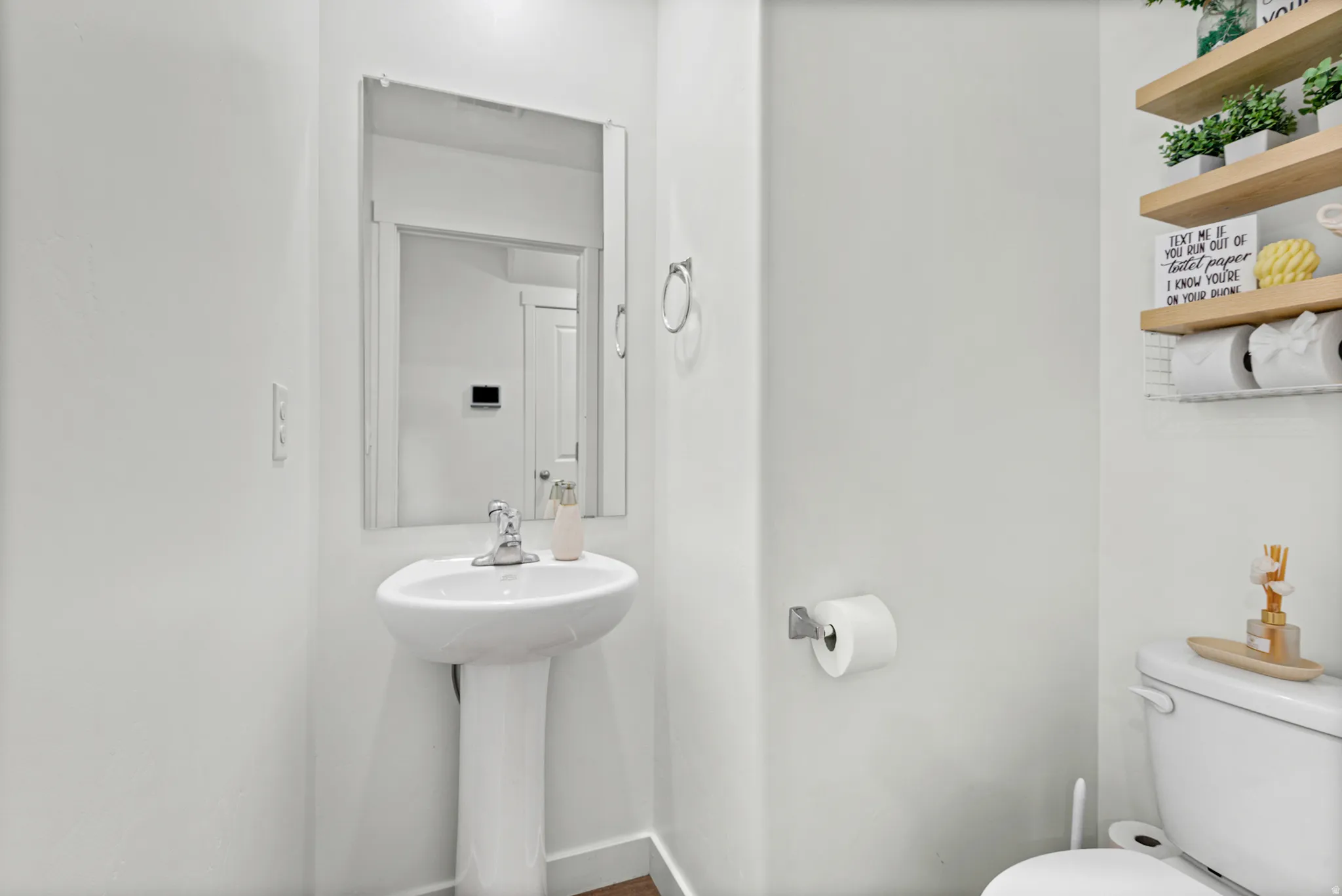 Pedestal sink with chrome faucet, wall-mounted mirror, and wood-finish floating shelves
