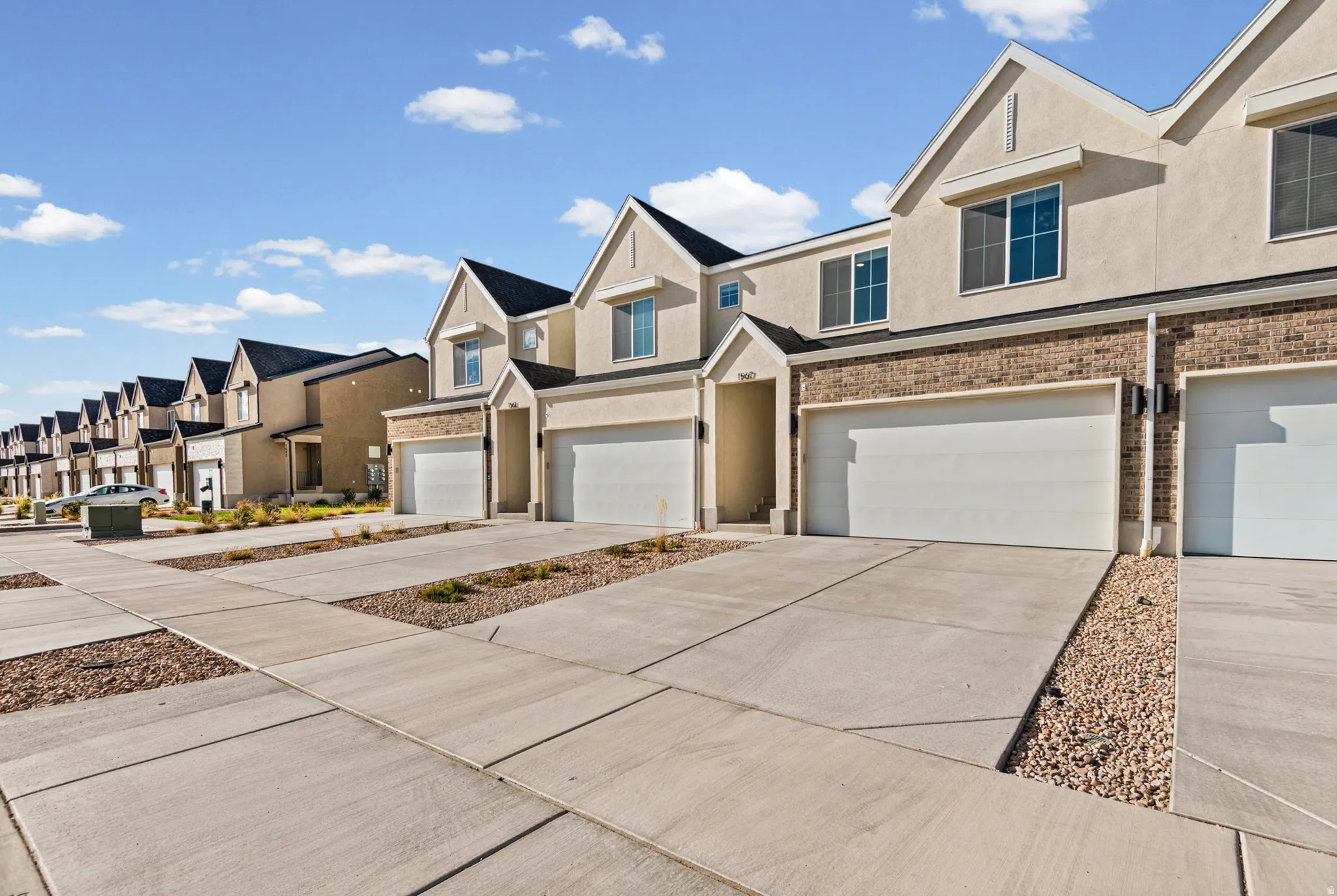 Contemporary townhome-style residences featuring stucco and brick exteriors, pitched shingle roofs, integrated garages, and concrete driveways