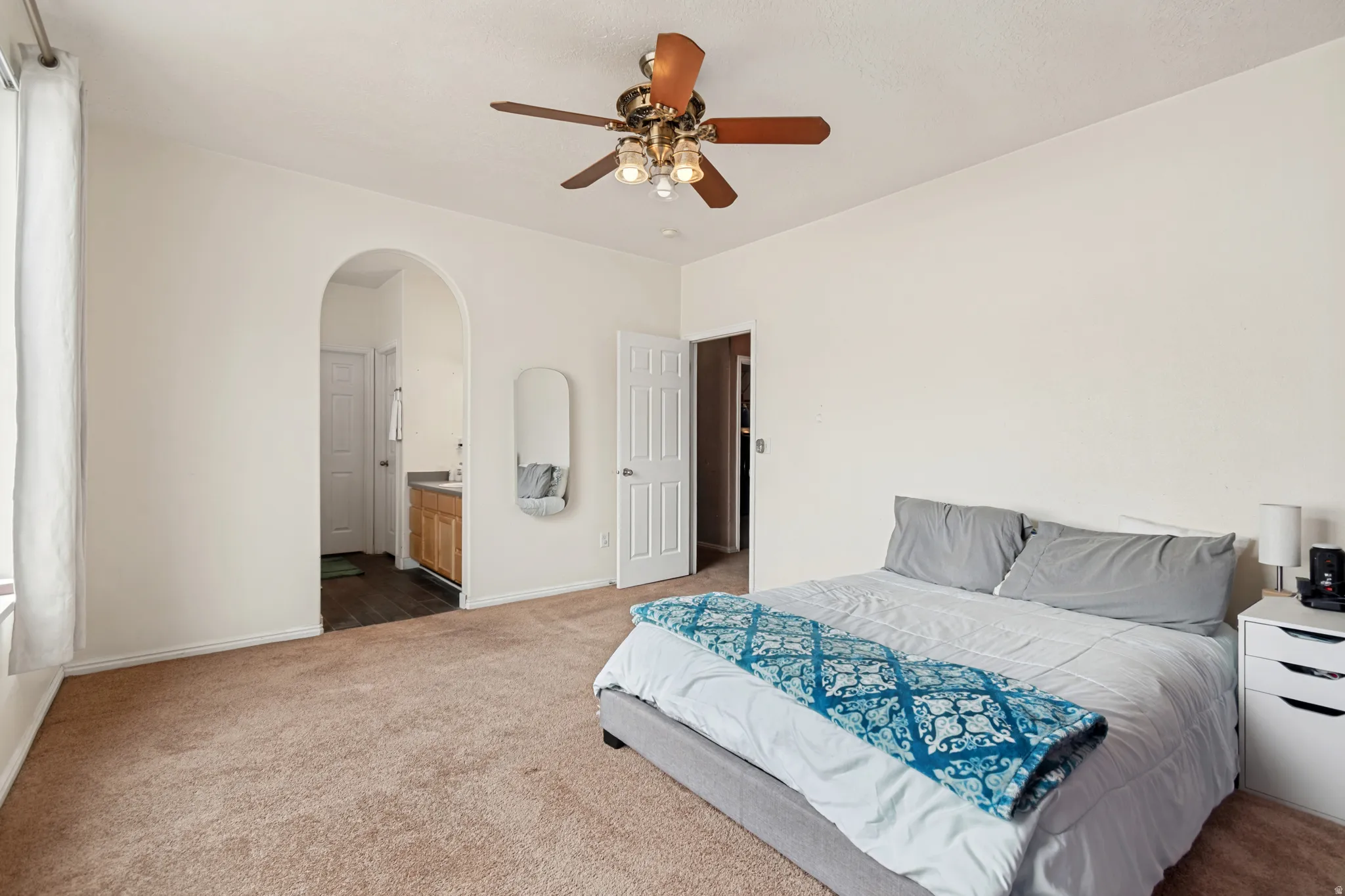 Spacious room featuring neutral carpeting, a ceiling fan with integrated lighting, and an arched doorway leading to an additional space with a vanity
