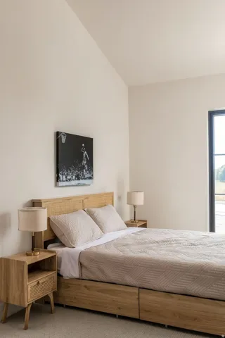 Vaulted ceiling, large window with dark trim, light wood-finish headboard, light wood-finish nightstands, light wood-finish bed frame with built-in drawers