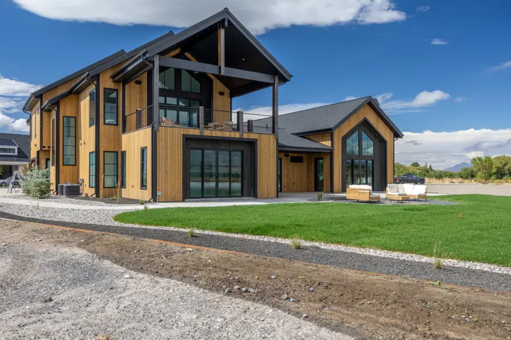 Modern exterior with light wood-finish siding, black-framed windows, a second-story balcony with metal railings, a dark gabled roof, and a green lawn