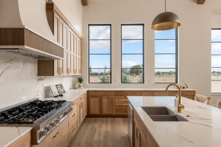 Vaulted ceilings with exposed beams, tall black-framed windows, wood-finish cabinetry, white countertops with natural veining, and a central island with an undermount sink