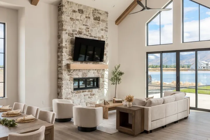Vaulted ceilings with exposed wood beams, stone-clad fireplace with wood mantel, built-in linear fireplace, expansive glass sliding doors, and large picture windows