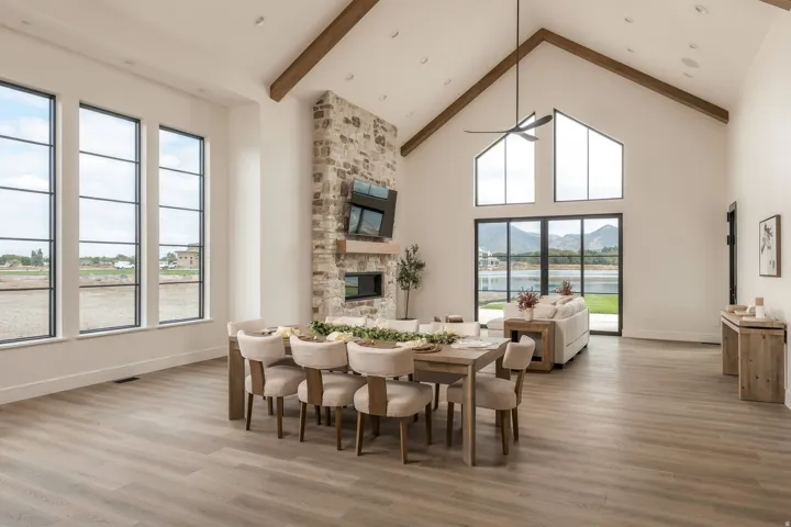 Vaulted ceilings with exposed beams, wood-finish flooring, a stone-clad fireplace with hearth, and extensive window panels providing abundant illumination
