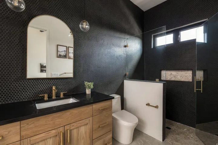 Black penny tile walls, natural wood-finish vanity, black countertop with undermount sink, brass finish fixtures, and a glass-enclosed shower