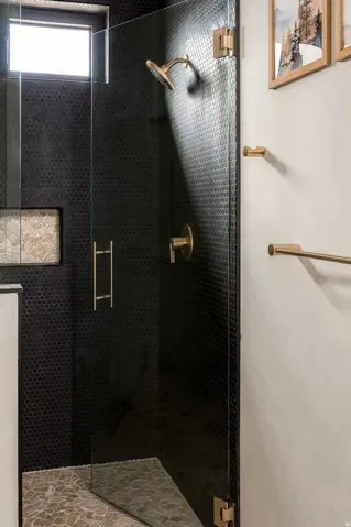 Walk-in shower featuring clear glass enclosure, black penny tile walls, gold-finish fixtures, and a transom window