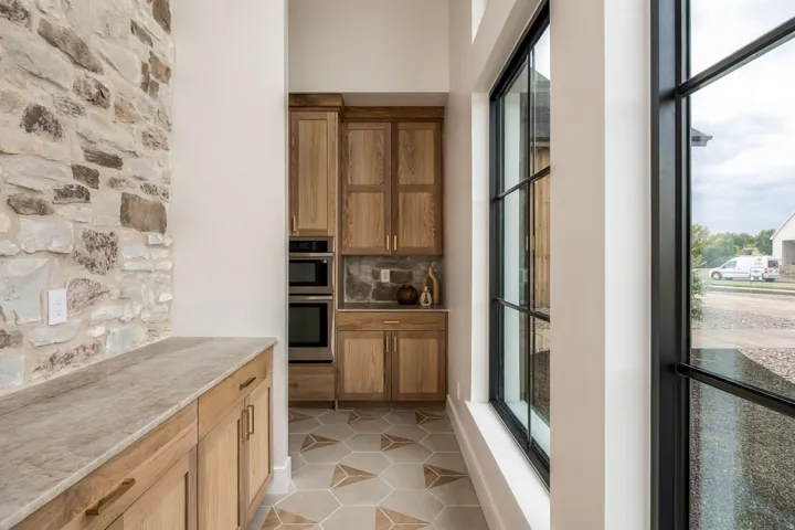 Stone accent wall, wood cabinetry with gold-tone hardware, built-in stainless steel appliances, patterned hexagonal floor tile, and black-framed windows