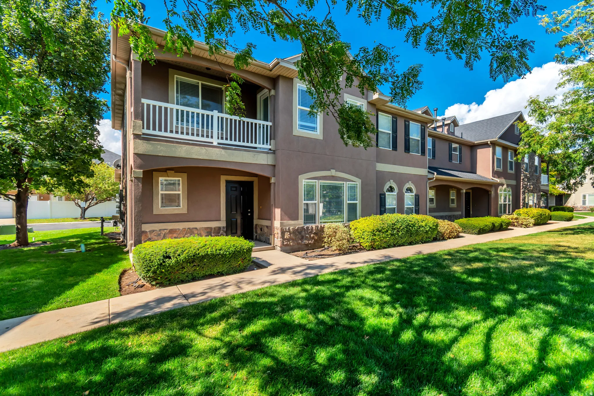 Stucco exterior with stone accent, upper-level balcony, ground-level entry, manicured shrubs, and a sidewalk