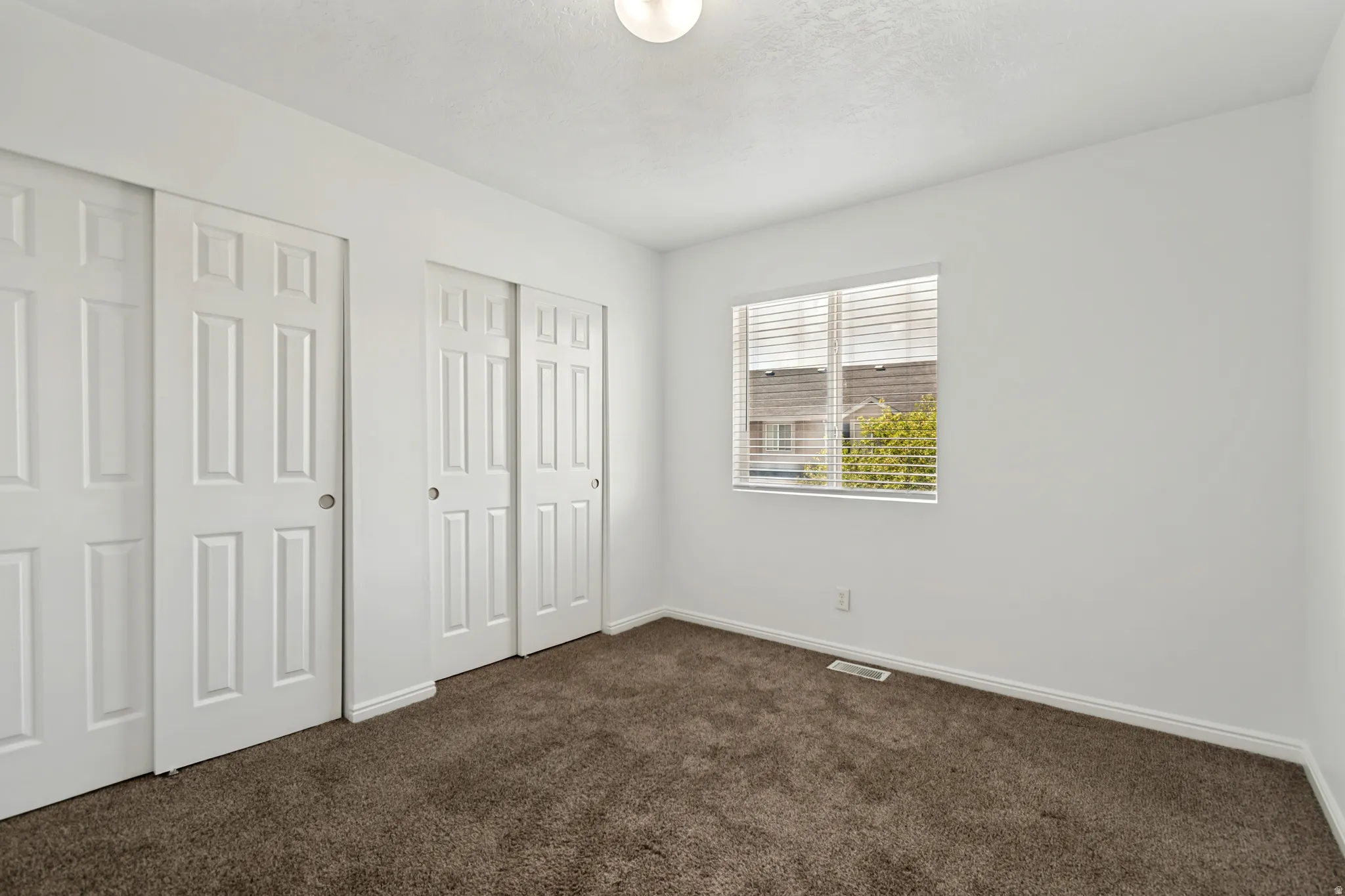 Neutral wall color, plush carpet, window with blinds, and two sets of bi-fold closet doors