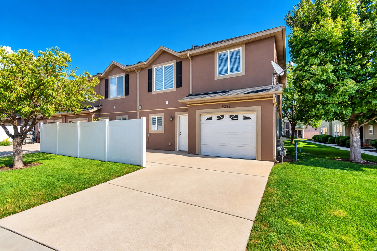 Tan exterior siding with white trim, a single-car garage, a concrete driveway, a white privacy fence, and manicured green lawns