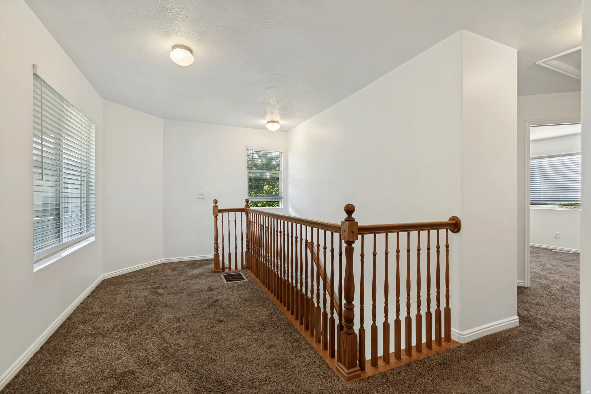 Carpeted landing with white walls, wood spindle railing, two surface-mounted light fixtures, and windows with horizontal blinds