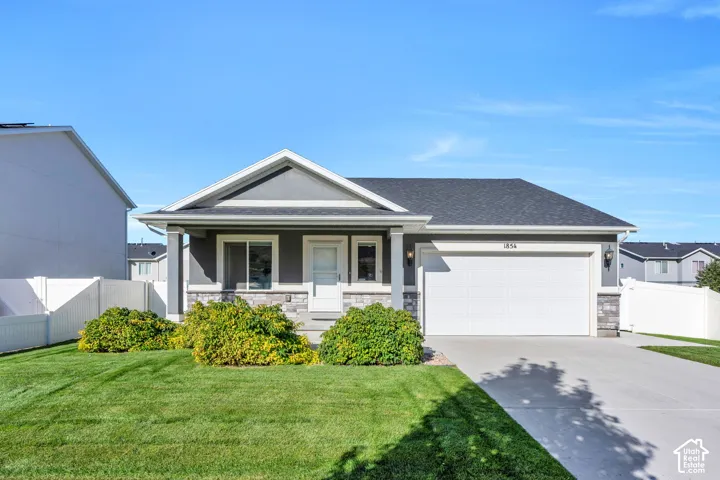 View of the front of the home. Modern ranch exterior featuring gray siding, a stone facade, an attached two car garage, a covered front porch, and a white privacy fence.