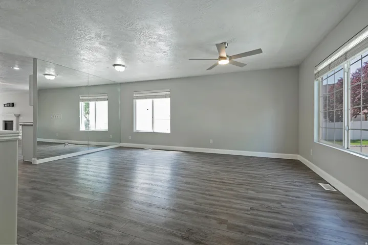 Spacious room featuring wood-finish flooring, a full-wall mirror, multiple windows, and a ceiling fan with integrated lighting