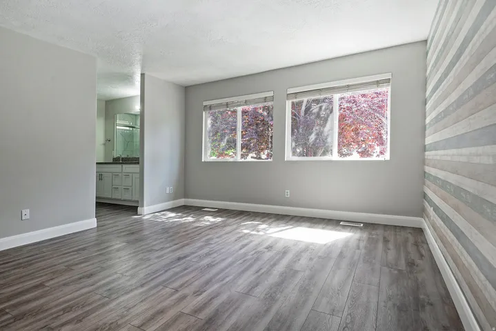 Wood-finish flooring, accent wall with horizontal paneling, two windows, and a partial bathroom with a dual vanity and mirror