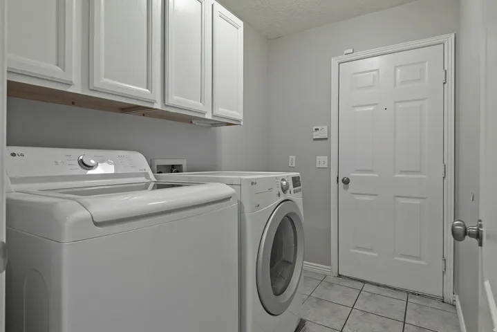 Integrated laundry area featuring white shaker-style cabinetry with a wood-finish shelf, light gray wall paint, and square floor tiles